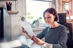 Woman wearing striped shirt sitting by the window at the table and reading mail in the office